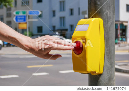 Close-up of a hand pressing a pedestrian crossing button at a signalized crosswalk in an urban environment. Concept of road safety, accessible transportation, traffic control and city infrastructure. Close-up of a hand pressing a pedestrian crossing button at a signalized crosswalk in an urban environment. Concept of road safety, accessible transportation, traffic control and city infrastructure. 141673074