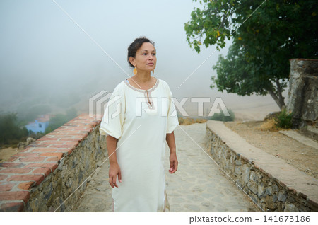 Woman in white traditional dress walking on a foggy stone path near a rustic wall Woman in white traditional dress walking on a foggy stone path near a rustic wall 141673186