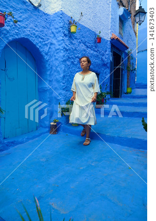 Woman walking through blue-washed streets of Chefchaouen in a white traditional dress Woman walking through blue-washed streets of Chefchaouen in a white traditional dress 141673403