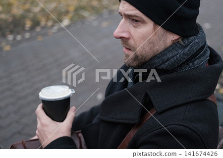 Bearded adult man holding hot drink paper cup in city bokeh. Serious male with coffee to go near residential building background. Morning commute story. Urban Lifestyle. Autumn Cold Weather Comfort Bearded adult man holding hot drink paper cup in city bokeh. Serious male with coffee to go near residential building background. Morning commute story. Urban Lifestyle. Autumn Cold Weather Comfort 141674426