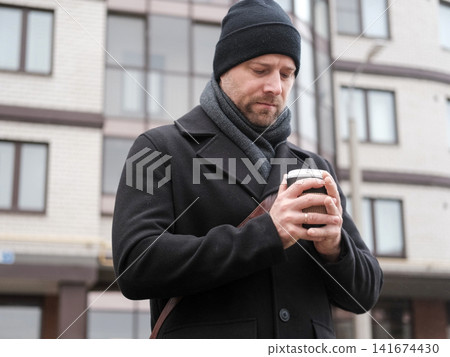 Bearded adult man holding hot drink paper cup in city bokeh. Serious male with coffee to go near residential building background. Morning commute story. Urban Lifestyle. Autumn Cold Weather Comfort Bearded adult man holding hot drink paper cup in city bokeh. Serious male with coffee to go near residential building background. Morning commute story. Urban Lifestyle. Autumn Cold Weather Comfort 141674430