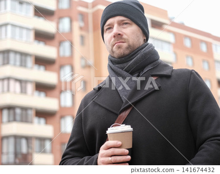Bearded adult man holding hot drink paper cup in city bokeh. Serious male with coffee to go near residential building background. Morning commute story. Urban Lifestyle. Autumn Cold Weather Comfort Bearded adult man holding hot drink paper cup in city bokeh. Serious male with coffee to go near residential building background. Morning commute story. Urban Lifestyle. Autumn Cold Weather Comfort 141674432
