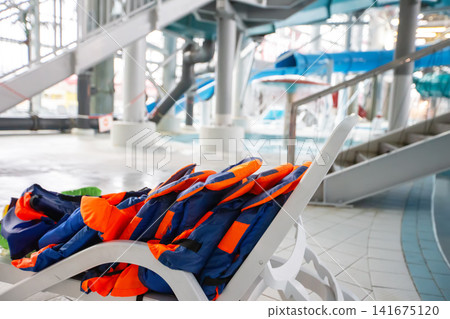 Colorful life jackets on a lounge chair in a bright indoor water park with water slides in the background Colorful life jackets on a lounge chair in a bright indoor water park with water slides in the background 141675120
