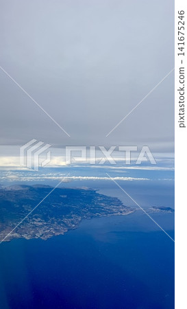 Aerial view of island coastline surrounded by deep blue ocean and layered clouds seen from airplane. Natural seascape with land, water and sky, travel and tourism concept, calm atmosphere, freedom and Aerial view of island coastline surrounded by deep blue ocean and layered clouds seen from airplane. Natural seascape with land, water and sky, travel and tourism concept, calm atmosphere, freedom and 141675246