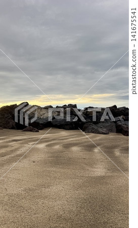 Natural rocky coastline with rippled sand patterns in foreground and dark volcanic stones under dramatic cloudy sunset sky, minimal seascape background for travel, nature, mindfulness, freedom and cal 141675541
