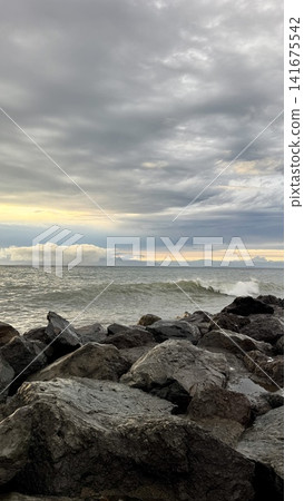 Powerful sea waves breaking against dark coastal rocks under dramatic cloudy sky at sunset. Natural ocean energy, rough water, and moody seascape suitable for travel, nature, weather, and coastal conc Powerful sea waves breaking against dark coastal rocks under dramatic cloudy sky at sunset. Natural ocean energy, rough water, and moody seascape suitable for travel, nature, weather, and coastal conc 141675542