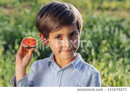 Smiling little boy holding freshly cut ripe fig slice while looking at camera. Healthy childhood Smiling little boy holding freshly cut ripe fig slice while looking at camera. Healthy childhood 141678872