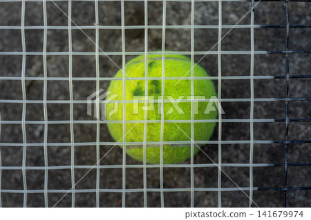 Neon green tennis ball pressed behind metal fence grid outdoors, close front view with blurred gravel background and vertical steel bars, showing sports training challenge and enclosure Neon green tennis ball pressed behind metal fence grid outdoors, close front view with blurred gravel background and vertical steel bars, showing sports training challenge and enclosure 141679974