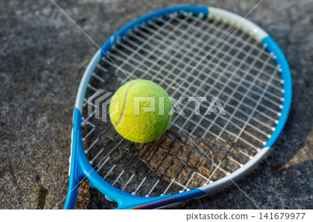 Blue tennis racket with yellow ball resting on rough outdoor pavement, close-up sports scene with shallow depth of field for fitness, recreation, competition, and training use Blue tennis racket with yellow ball resting on rough outdoor pavement, close-up sports scene with shallow depth of field for fitness, recreation, competition, and training use 141679977