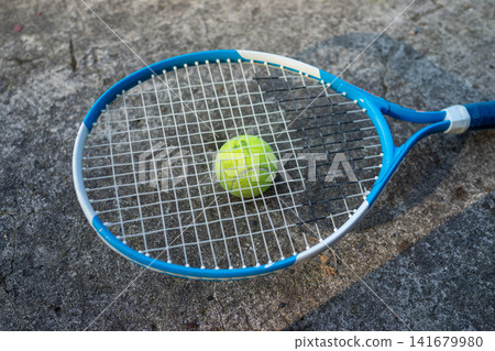 Blue tennis racket with yellow green tennis ball resting on concrete pavement, outdoor sports equipment scene for training, practice, recreation, and active play in natural daylight Blue tennis racket with yellow green tennis ball resting on concrete pavement, outdoor sports equipment scene for training, practice, recreation, and active play in natural daylight 141679980