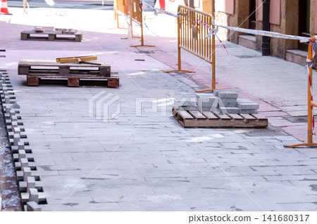 Unfinished section of new stone tile pavement. Wooden pallets with stacked building materials and orange metal safety barriers. Sidewalk renovation and paving works on a city street Unfinished section of new stone tile pavement. Wooden pallets with stacked building materials and orange metal safety barriers. Sidewalk renovation and paving works on a city street 141680317