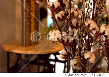 Natural cotton bolls arranged in a glass vase inside a flower shop. Soft white fibers contrast with brown dried seed pods and decorative pussy willow branches Natural cotton bolls arranged in a glass vase inside a flower shop. Soft white fibers contrast with brown dried seed pods and decorative pussy willow branches 141680335