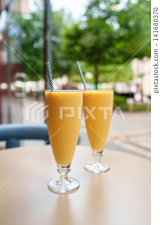 Two fresh banana smoothies served in tall glasses with paper straws on a table inside a cozy cafeteria. Creamy tropical beverages against a softly blurred street view through the cafe window Two fresh banana smoothies served in tall glasses with paper straws on a table inside a cozy cafeteria. Creamy tropical beverages against a softly blurred street view through the cafe window 141680370