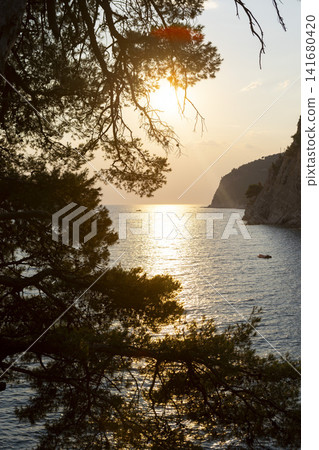 Sunset glow over tranquil coastal bay framed by pine branches, rugged headland, rocky shoreline, and rippled water, creating peaceful seascape with warm reflections and secluded cove Sunset glow over tranquil coastal bay framed by pine branches, rugged headland, rocky shoreline, and rippled water, creating peaceful seascape with warm reflections and secluded cove 141680420