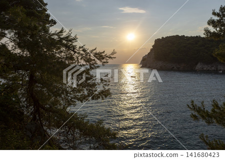Sunset glow over tranquil coastal bay framed by pine branches, rugged headland, rocky shoreline, and rippled water, creating peaceful seascape with warm reflections and secluded cove Sunset glow over tranquil coastal bay framed by pine branches, rugged headland, rocky shoreline, and rippled water, creating peaceful seascape with warm reflections and secluded cove 141680423