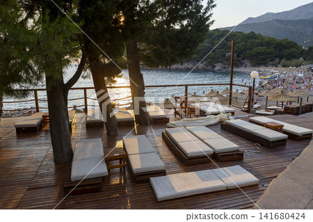 Sunlit beach lounge chairs and wicker umbrellas on wooden deck overlooking crowded pebble shore, warm late afternoon light casting long shadows across seaside resort setting Sunlit beach lounge chairs and wicker umbrellas on wooden deck overlooking crowded pebble shore, warm late afternoon light casting long shadows across seaside resort setting 141680424