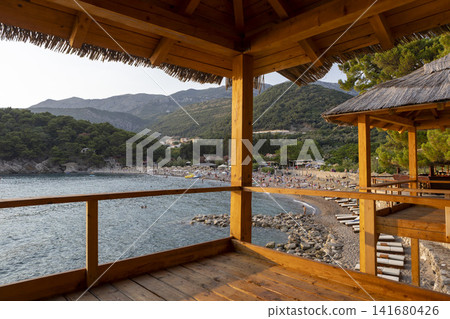 Wooden seaside pavilion with shingled roof and railing overlooking rocky cove, calm blue water, distant hills, and warm golden hour light across coastal floorboards Wooden seaside pavilion with shingled roof and railing overlooking rocky cove, calm blue water, distant hills, and warm golden hour light across coastal floorboards 141680426