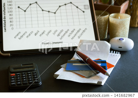 Business workspace representing financial planning, data analysis, and productivity with a tablet displaying a graph, a calculator, credit cards, a pen, and receipts on a dark surface Business workspace representing financial planning, data analysis, and productivity with a tablet displaying a graph, a calculator, credit cards, a pen, and receipts on a dark surface 141682967