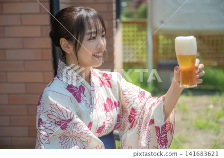 一位身穿浴衣的女子舉起啤酒,帶著微笑敬酒。 一位身穿浴衣的女子舉起啤酒,帶著微笑敬酒。 141683621