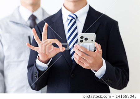 A young man in a business suit and a middle-aged man in work clothes are making a circular motion while holding their smartphones. A young man in a business suit and a middle-aged man in work clothes are making a circular motion while holding their smartphones. 141685045