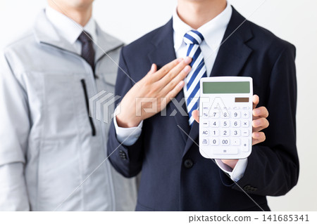 一位身穿西裝的年輕男子和一位身穿工作服的中年男子,雙手放在胸前,手裡拿著一台白色計算器。 一位身穿西裝的年輕男子和一位身穿工作服的中年男子,雙手放在胸前,手裡拿著一台白色計算器。 141685341