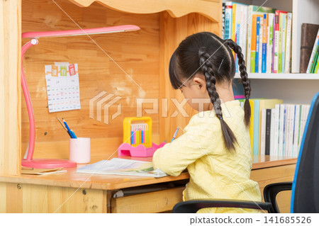 Asian little girl studying at wooden desk, back view child doing homework at home, cozy study environment concept 141685526