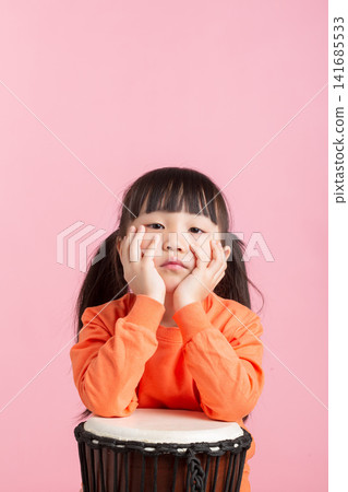 Cute Asian little girl playing drum, smiling child with percussion instrument on pink background, music education concept Cute Asian little girl playing drum, smiling child with percussion instrument on pink background, music education concept 141685533