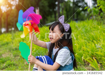 Happy Young East Asian Girl with Purple Bow Sitting in Sunny Green Meadow Holding Colorful Rainbow Pinwheel with Joyful Smile and Bright Summer Vibes. Happy Young East Asian Girl with Purple Bow Sitting in Sunny Green Meadow Holding Colorful Rainbow Pinwheel with Joyful Smile and Bright Summer Vibes. 141685542