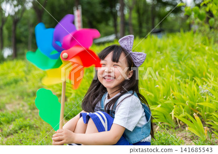 Happy Young East Asian Girl with Purple Bow Sitting in Sunny Green Meadow Holding Colorful Rainbow Pinwheel with Joyful Smile and Bright Summer Vibes. Happy Young East Asian Girl with Purple Bow Sitting in Sunny Green Meadow Holding Colorful Rainbow Pinwheel with Joyful Smile and Bright Summer Vibes. 141685543