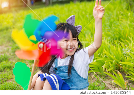 Happy Young East Asian Girl with Purple Bow Sitting in Sunny Green Meadow Holding Colorful Rainbow Pinwheel with Joyful Smile and Bright Summer Vibes. 141685544