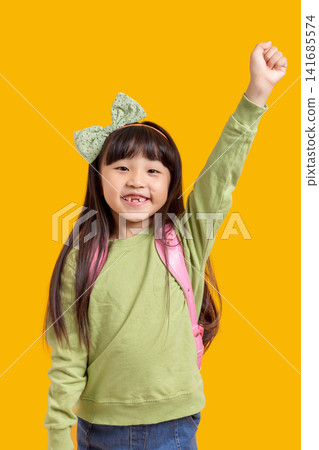 A happy Asian schoolgirl raises her hand with her backpack on; against a yellow background, the excited child exudes a sense of success and confidence. A happy Asian schoolgirl raises her hand with her backpack on; against a yellow background, the excited child exudes a sense of success and confidence. 141685574