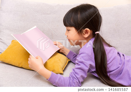 Focused Asian little girl reading book on sofa at home, early learning and literacy concept 141685587