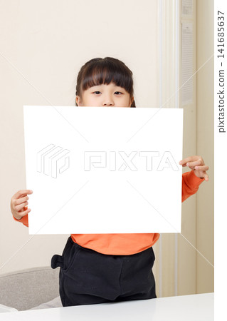 Young girl standing in a bright room holding an empty white card under her face, looking directly at the camera. Young girl standing in a bright room holding an empty white card under her face, looking directly at the camera. 141685637