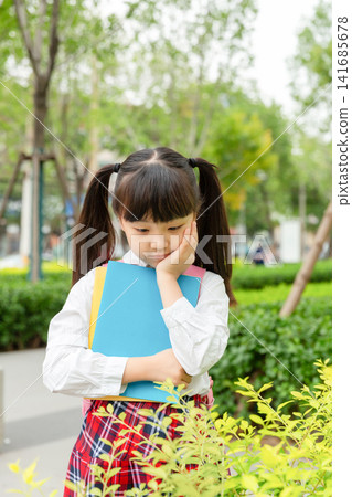 A thoughtful young student holding a blue folder and looking down while standing next to green bushes. A thoughtful young student holding a blue folder and looking down while standing next to green bushes. 141685678