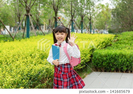 A cheerful elementary school girl in a plaid uniform walks along a green garden path with a pink backpack. 141685679