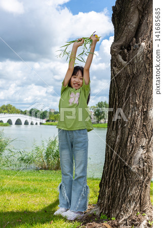 A cheerful young girl leaning against a tree trunk and raising a willow branch high in a sunny park. 141685685