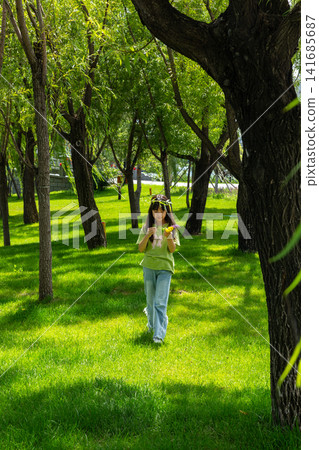 A young girl wearing a leaf crown stands amidst tall green trees on a sunlit grassy field. A young girl wearing a leaf crown stands amidst tall green trees on a sunlit grassy field. 141685687