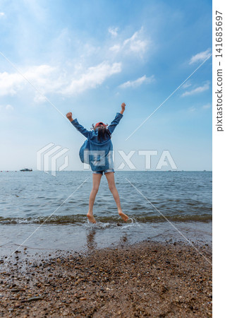 A carefree young girl in a red cap jumps high into the air on a sunny beach facing the ocean. A carefree young girl in a red cap jumps high into the air on a sunny beach facing the ocean. 141685697