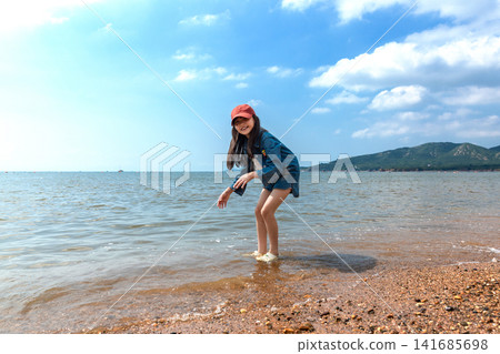 A young girl sitting on the seashore and splashing seawater with her hands on a bright summer day. A young girl sitting on the seashore and splashing seawater with her hands on a bright summer day. 141685698