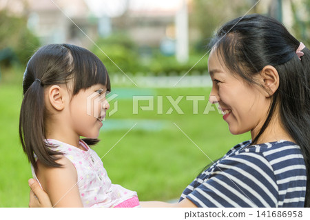 A happy mother and her little daughter smiling and looking at each other affectionately in a green park during summertime. A happy mother and her little daughter smiling and looking at each other affectionately in a green park during summertime. 141686958