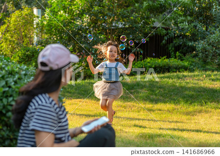 Little girl playing with bubbles in park while parent using smartphone, modern parenting and screen distraction concept Little girl playing with bubbles in park while parent using smartphone, modern parenting and screen distraction concept 141686966