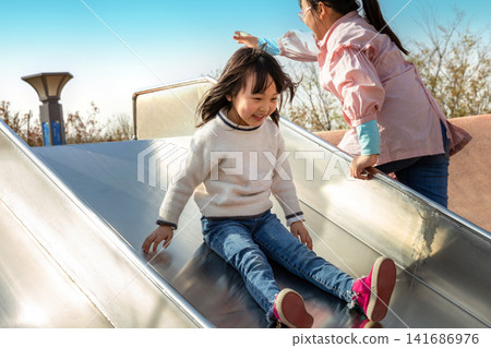 Excited little girl sliding down a metal slide at the playground on a sunny day Excited little girl sliding down a metal slide at the playground on a sunny day 141686976