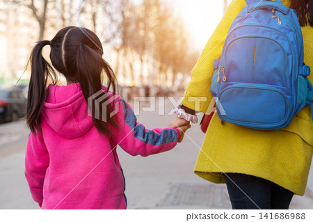A mother carrying a schoolbag and walking hand-in-hand with her daughter on their way home from school; close-up of their backs. 141686988