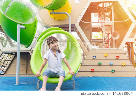 A young girl in a white t-shirt and jeans sits inside a green tunnel at a modern outdoor playground with warm sunlight. A young girl in a white t-shirt and jeans sits inside a green tunnel at a modern outdoor playground with warm sunlight. 141688064