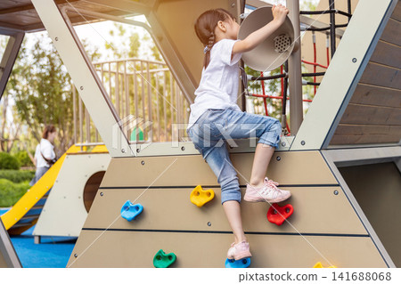 A determined little girl uses colorful holds to climb a wooden bouldering structure at an outdoor recreation area. A determined little girl uses colorful holds to climb a wooden bouldering structure at an outdoor recreation area. 141688068