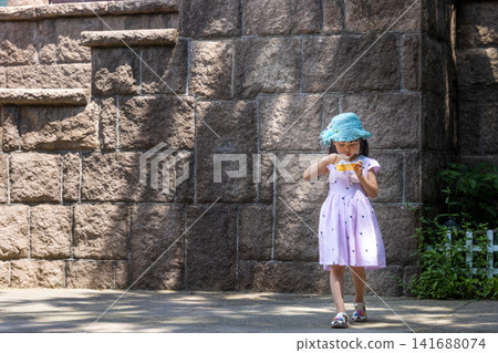 A little girl walks along a rustic stone wall in the park while eating from a yellow bowl on a sunny afternoon A little girl walks along a rustic stone wall in the park while eating from a yellow bowl on a sunny afternoon 141688074