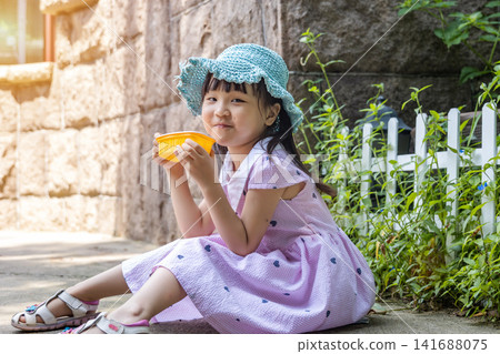 A charming Asian girl in a pink dress and straw hat sits on the ground and smiles while enjoying a snack in the park. A charming Asian girl in a pink dress and straw hat sits on the ground and smiles while enjoying a snack in the park. 141688075