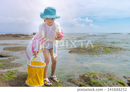 Little girl standing on a beach covered with bright green sea moss while holding a yellow bucket. Little girl standing on a beach covered with bright green sea moss while holding a yellow bucket. 141688252