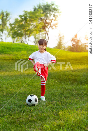 Smiling young girl dribbling a classic soccer ball on a lush green lawn surrounded by trees. Smiling young girl dribbling a classic soccer ball on a lush green lawn surrounded by trees. 141688257