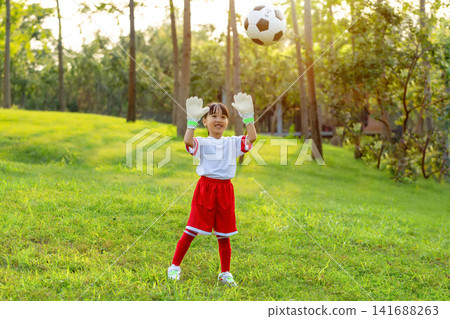 Enthusiastic young goalkeeper wearing gloves and catching a soccer ball in mid-air at the park. Enthusiastic young goalkeeper wearing gloves and catching a soccer ball in mid-air at the park. 141688263